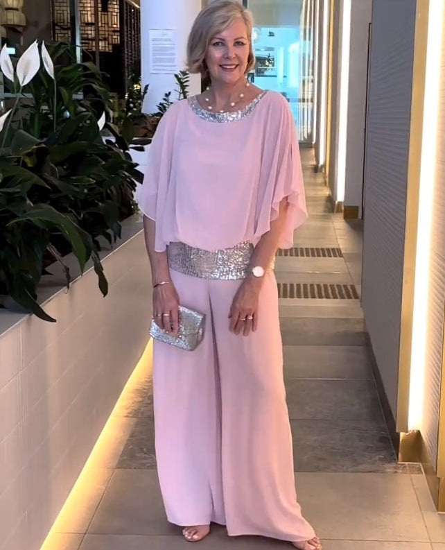 Woman in a pink elegant set with shimmering belt and neckline, holding a silver clutch. Indoor setting with plants.