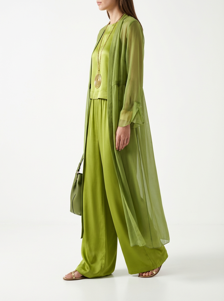Side view of model wearing olive green three-piece set with sheer duster and flowing pants, paired with gold necklace and carrying matching green bag in studio setting.