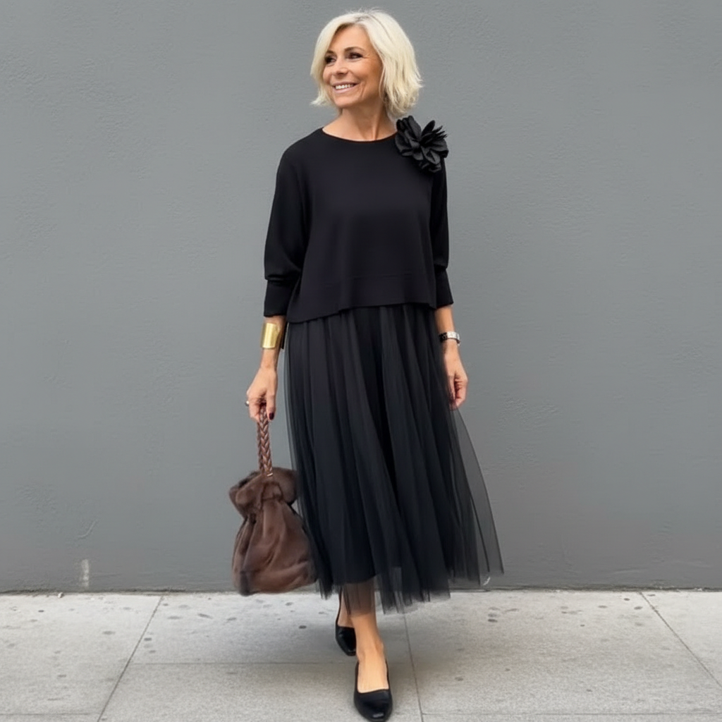 Stylish woman in black outfit with pleated skirt and large flower detail, holding brown bag, outdoor background.