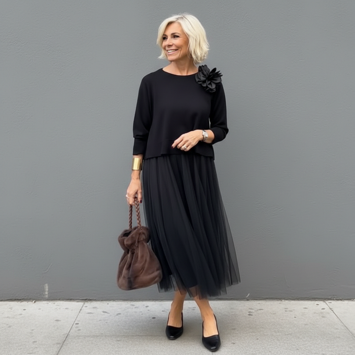 Woman in black pleated skirt outfit with flower detail, carrying brown bag, outside setting.