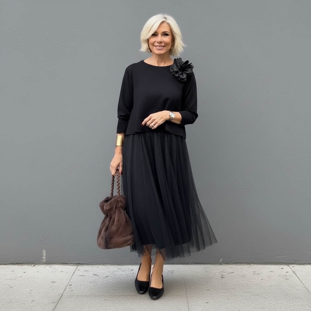 Sophisticated woman in black outfit with pleated skirt and flower detail, holding brown bag, urban setting.