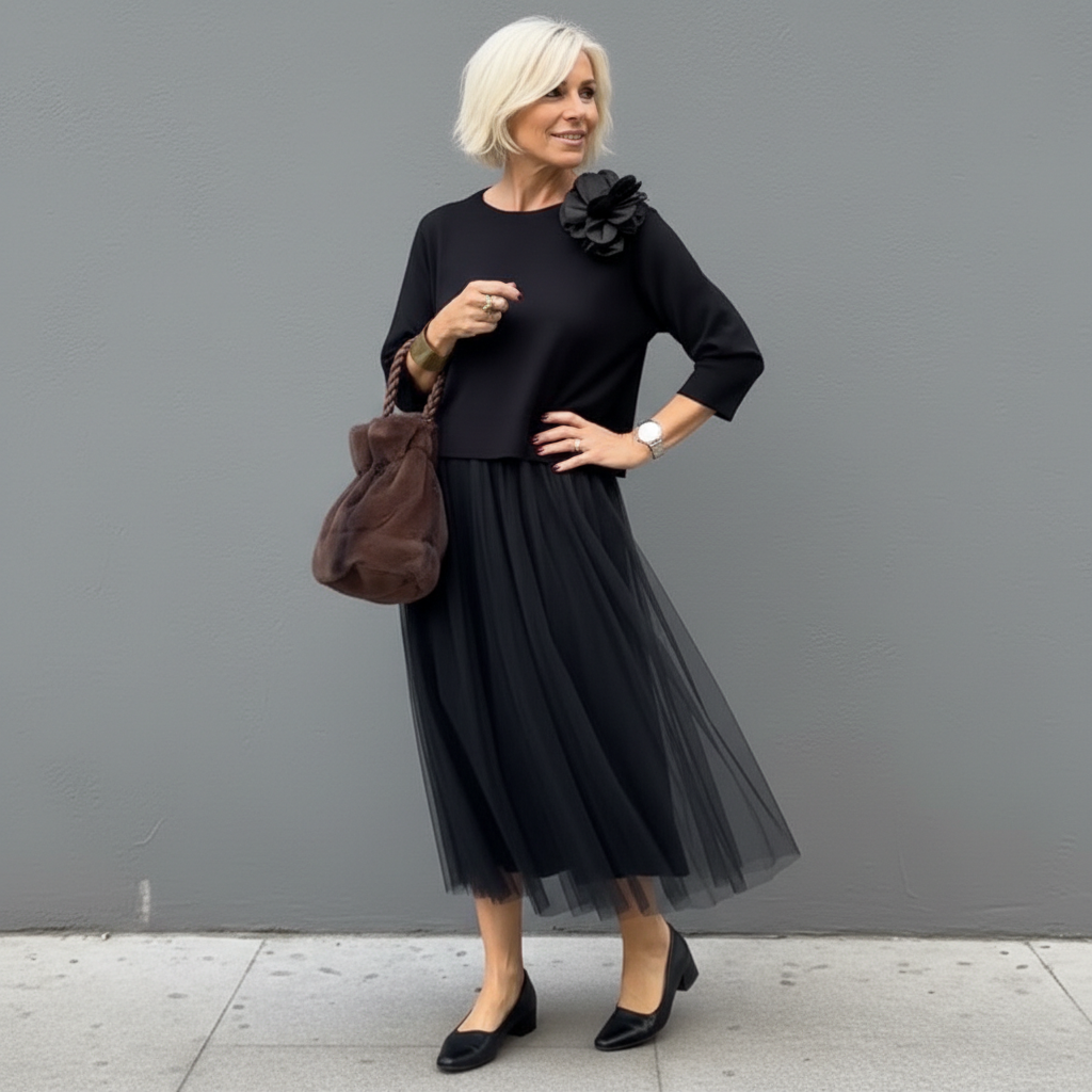Elegant woman in black outfit with pleated skirt and flower detail, by a gray wall.