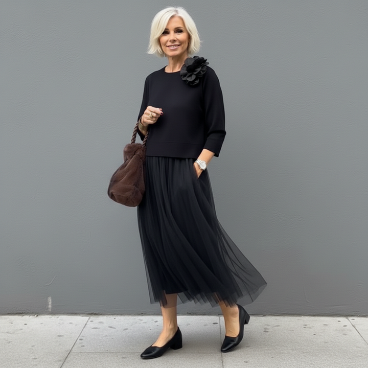 Elegant woman in black outfit with pleated skirt, flower detail and brown handbag, standing outside.