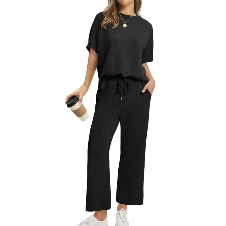 Woman in a sleek black set with loose top and drawstring pants, holding a coffee, in a studio setting.