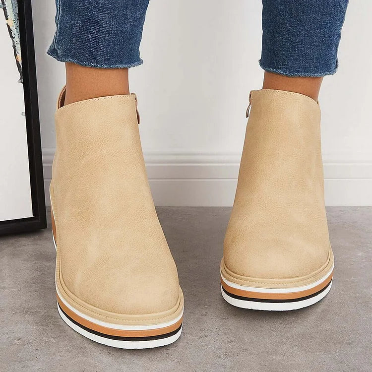 Front view of beige luxe boots with wedge soles, worn with frayed jeans, on a gray floor.