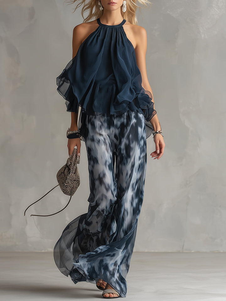 Elegant halter neck top with flowing layers paired with wide-leg patterned pants and a delicate handbag. Studio setting.