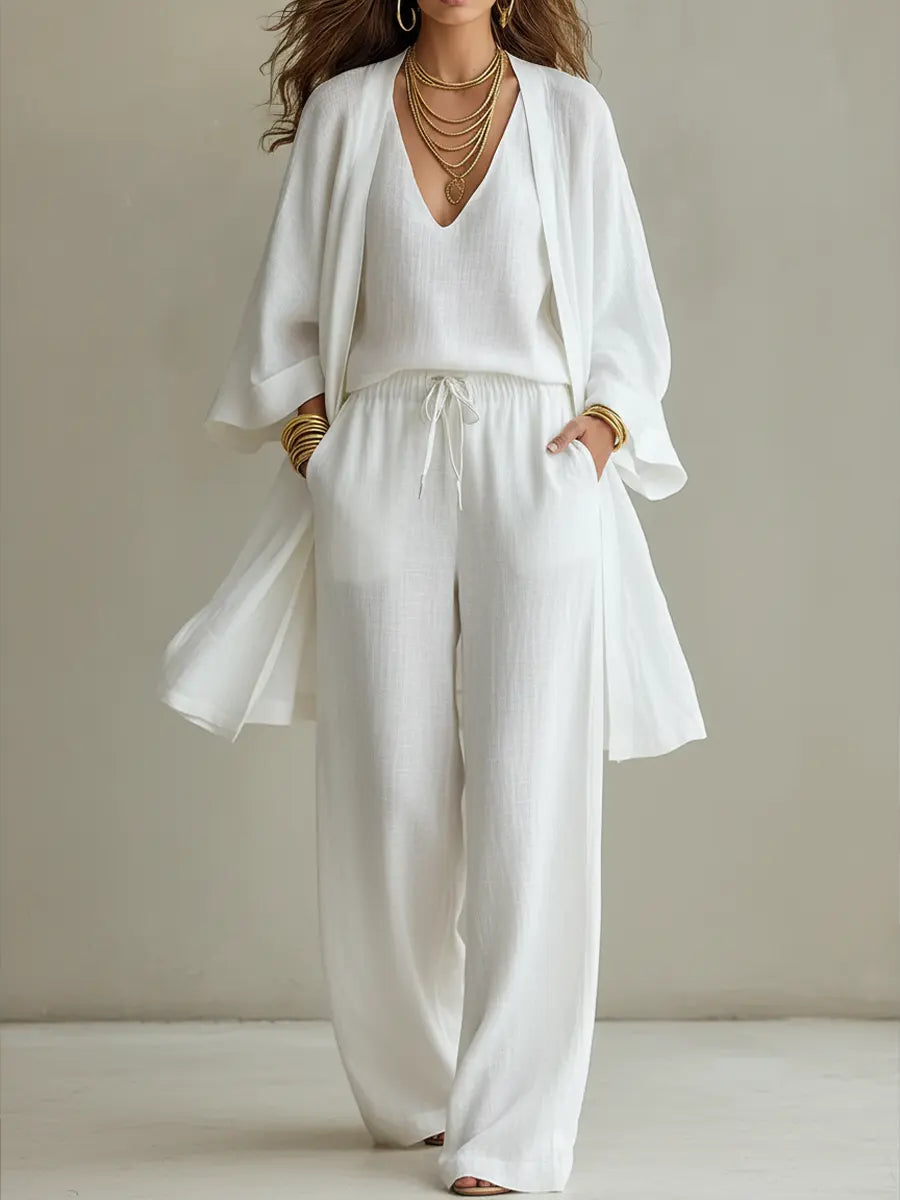 White three-piece relaxed homewear set with v-neck top, drawstring pants, and flowy overcoat in a studio setting.