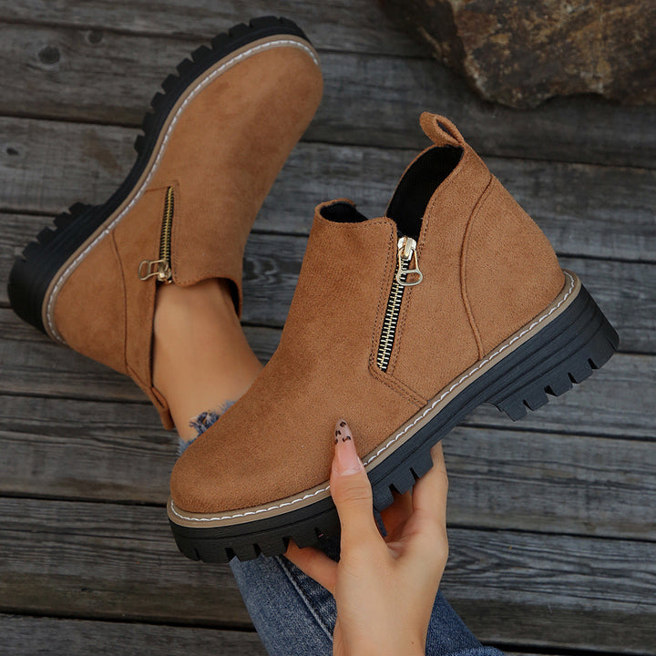 Brown ankle boots with zipper, held in hand on a rustic wooden surface.