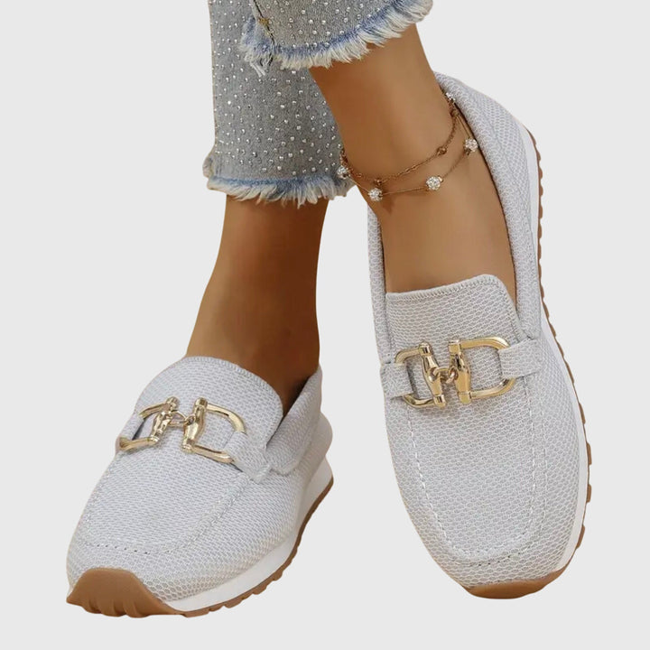 Light gray orthopedic loafers with gold details worn with frayed jeans and anklet on a light background.