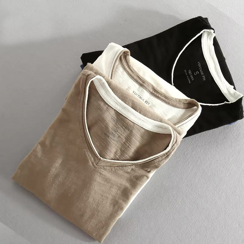 Stack of folded shirts in beige, white, and black with contrast trim, on a gray surface.