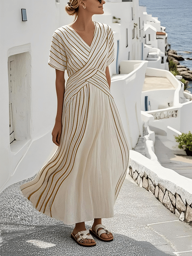 Elegant loose striped maxi dress in white with gold stripes, worn outdoors in a coastal, white building-lined street.