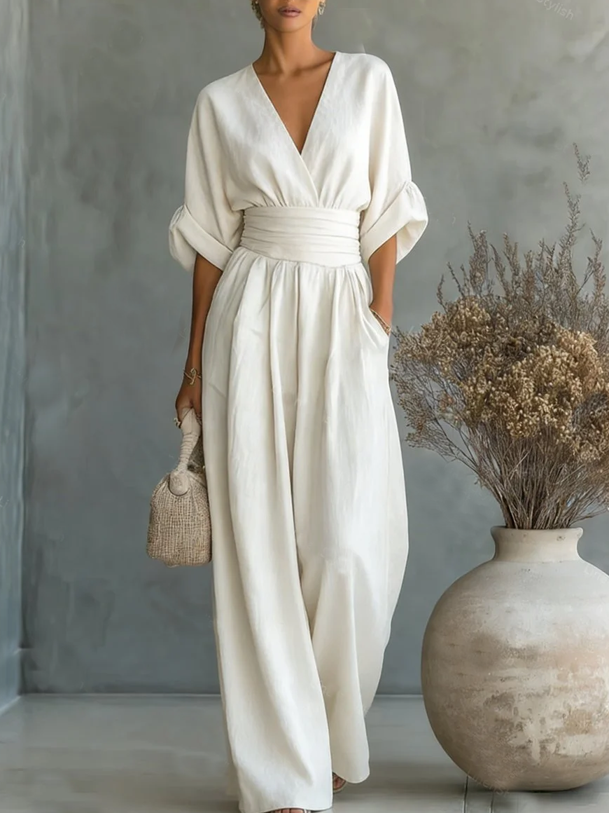 White V-neck high-waist jumpsuit featuring rolled-up sleeves, set in a minimalist studio with a large vase and dried flowers.
