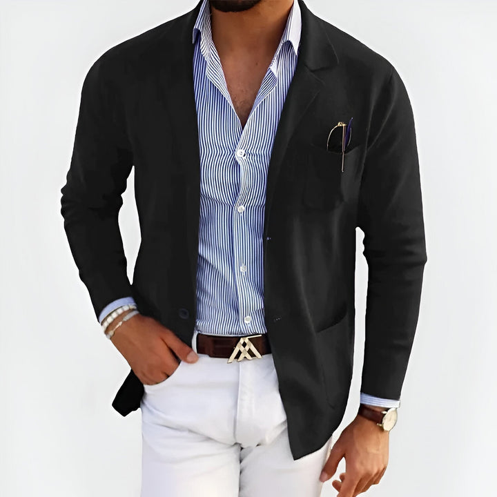 Man wearing a black lightweight jacket over a striped shirt, paired with white pants, in a studio setting.