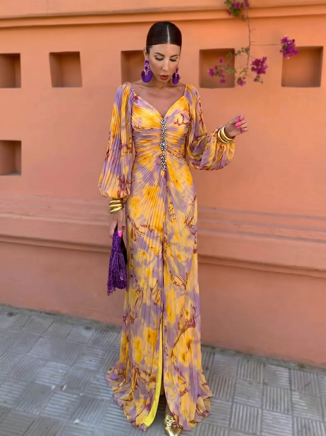 Woman in a vibrant long floral dress with a deep neckline, standing against a warm orange wall with purple flowers.