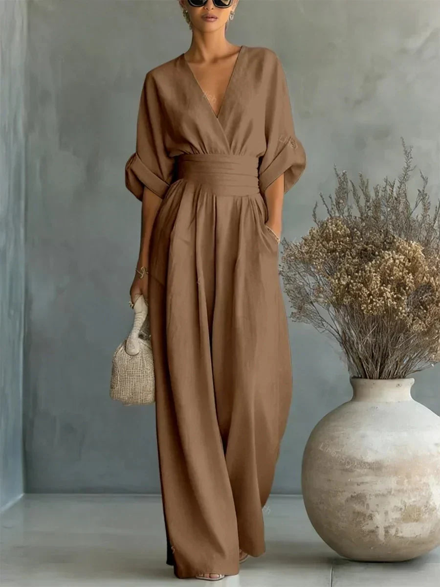 Brown V-neck high-waist jumpsuit with rolled-up sleeves, styled in a studio with earthy tones and a large vase.