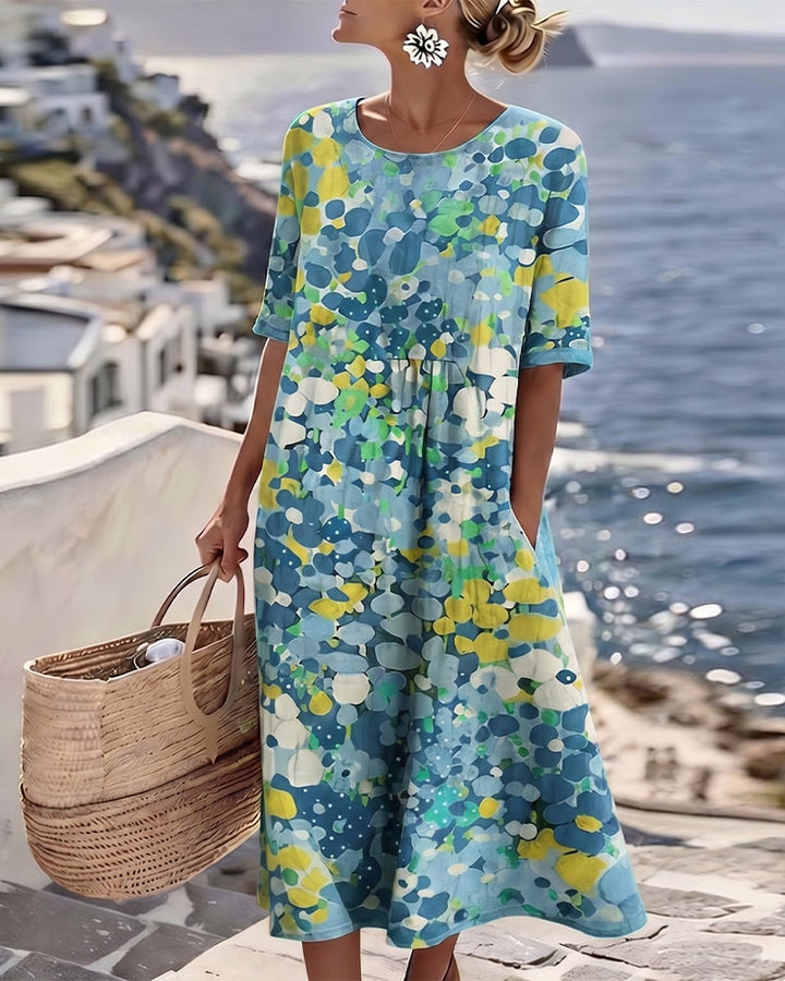 Woman in blue floral midi dress, holding wicker bag on sunny coastal terrace with ocean view.
