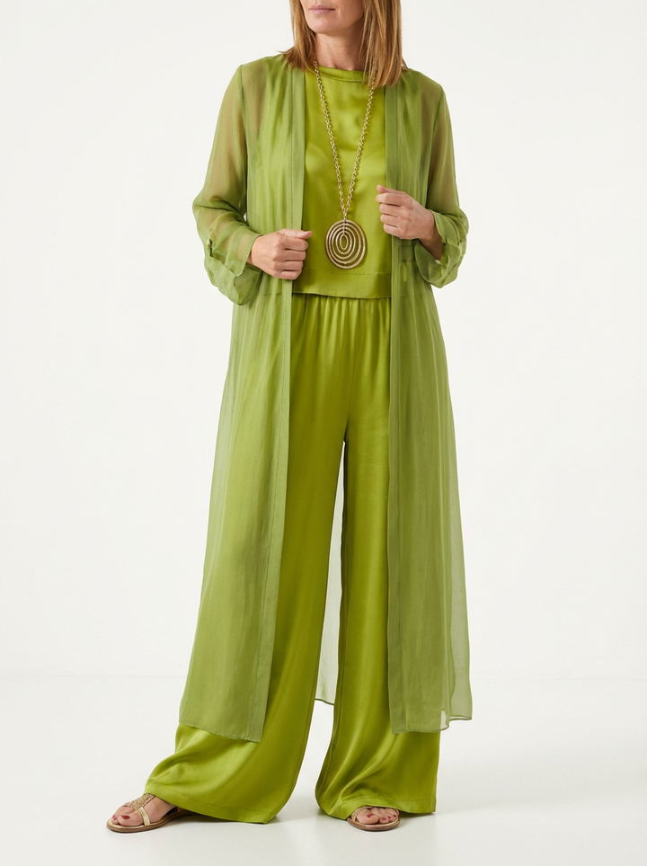 Model wearing olive green three-piece set featuring a sheer long duster, sleeveless top, wide-leg pants, gold necklace, and sandals in minimal studio background.