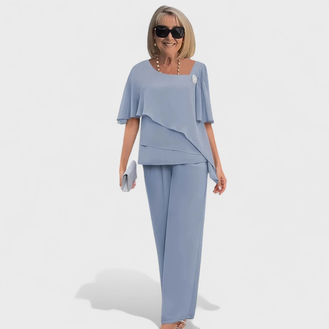 Woman wearing a light blue elegant two-piece set with layered top and wide-leg pants, holding a small clutch in a studio setting.