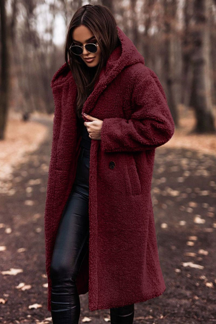 Woman in burgundy winter jacket and sunglasses walking on a leaf-strewn path in a wooded area.
