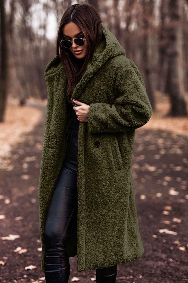 Woman in olive green winter jacket and sunglasses walking on a leaf-strewn path in a wooded area.