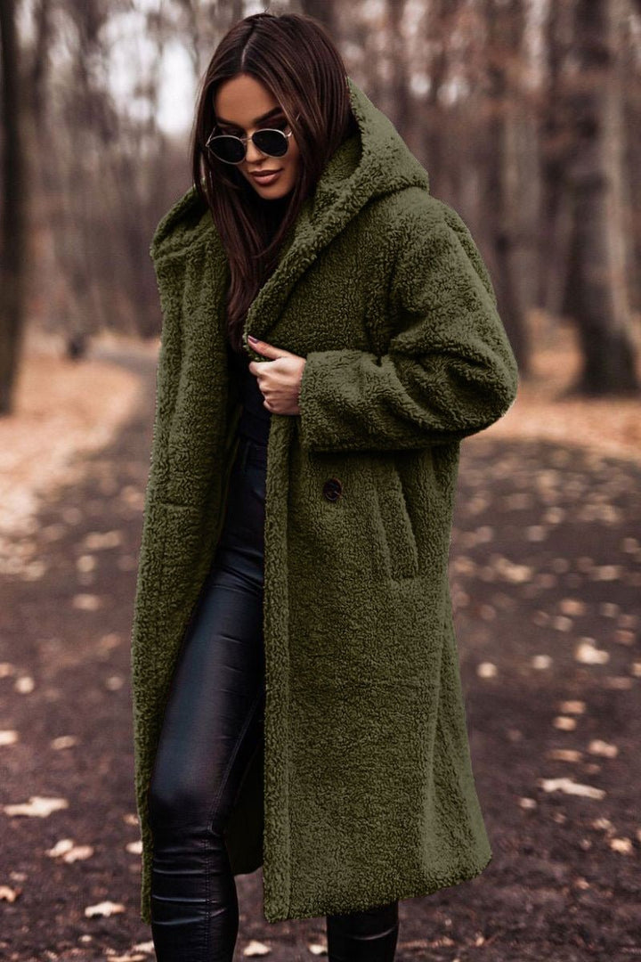 Woman in olive green winter jacket and sunglasses walking on a leaf-strewn path in a wooded area.
