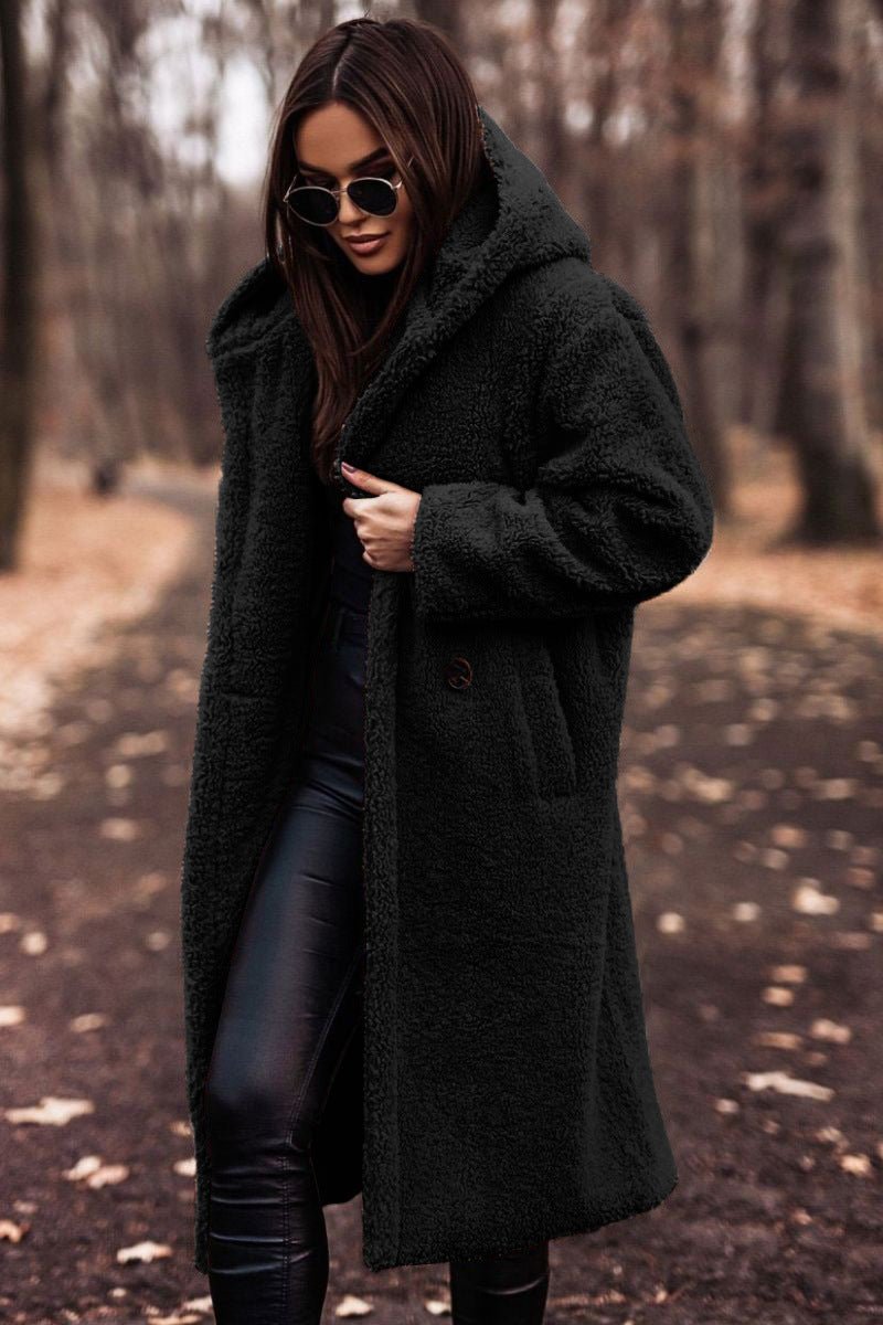 Woman in black winter jacket and sunglasses walking on a leaf-strewn path in a wooded area.