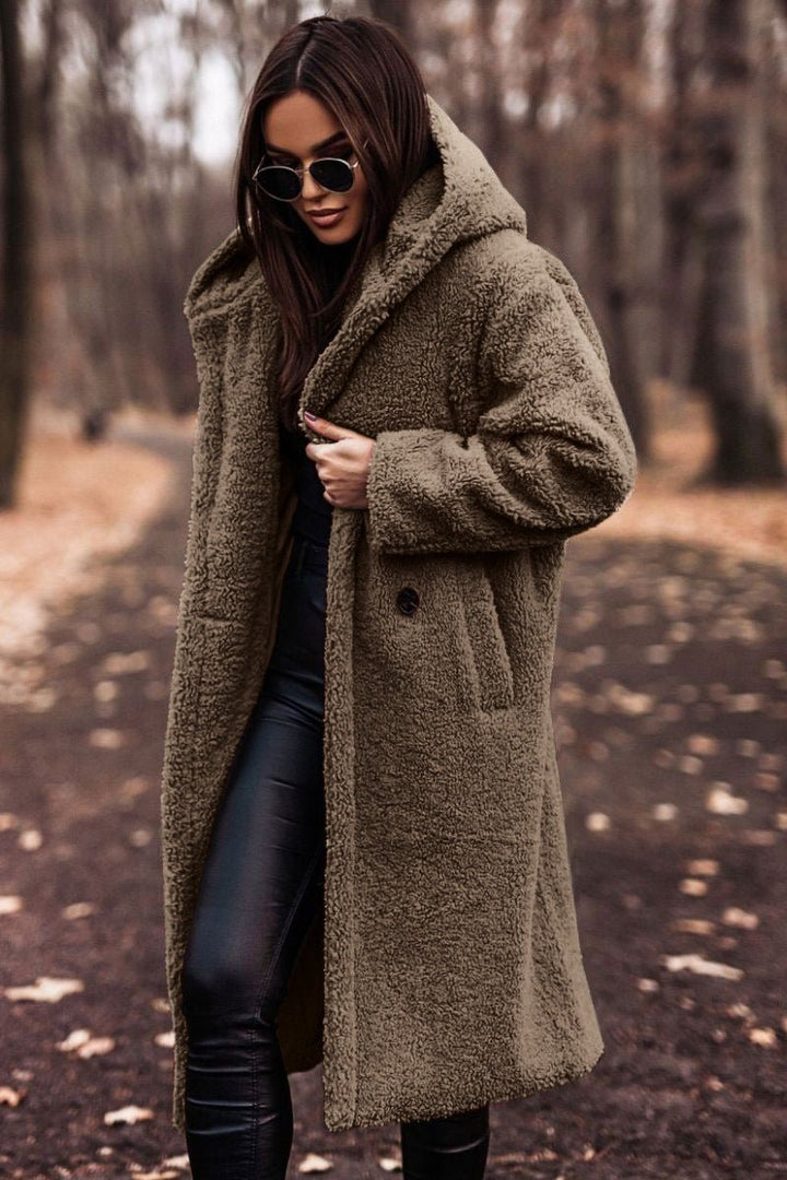 Woman in taupe winter jacket and sunglasses walking on a leaf-strewn path in a wooded area.