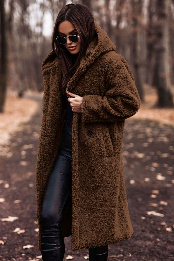 Woman in brown winter jacket and sunglasses walking on a leaf-strewn path in a wooded area.