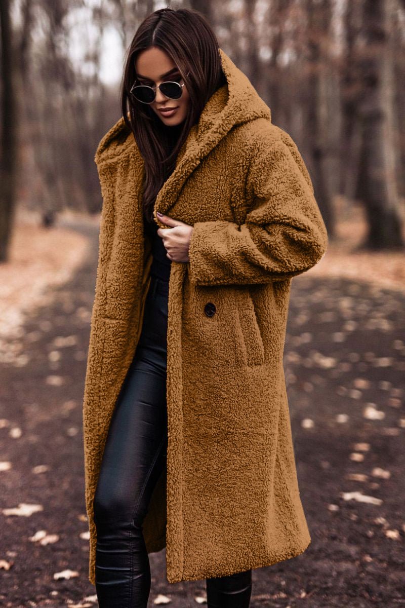 Woman in tan winter jacket and sunglasses walking on a leaf-strewn path in a wooded area.