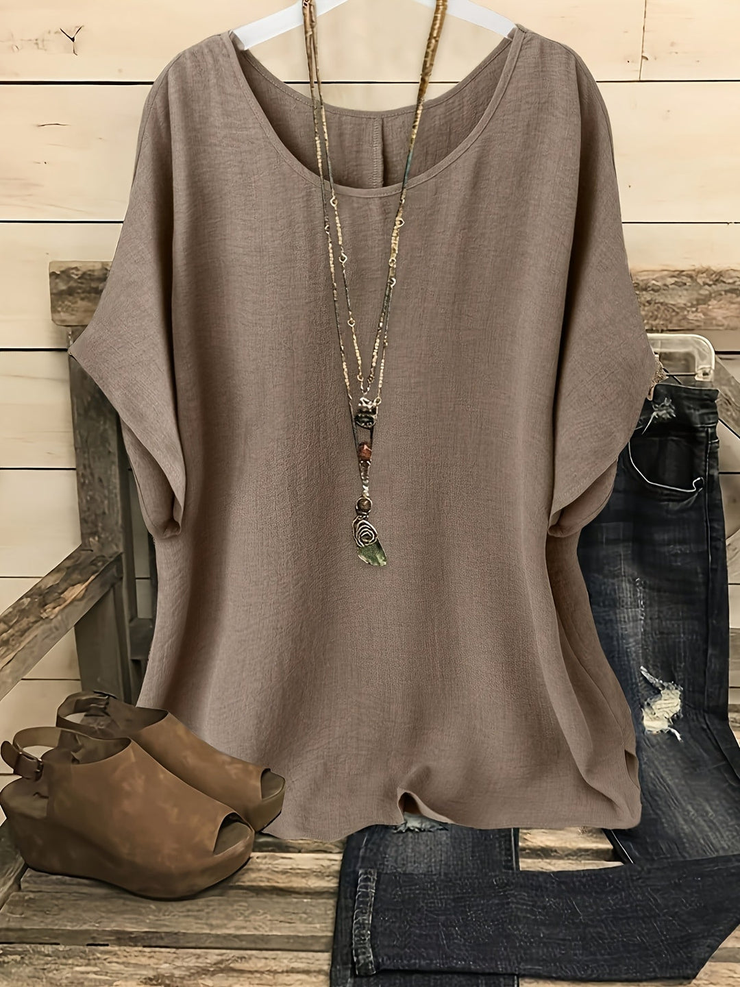Beige loose summer top on hanger with necklace, sandals, and jeans on rustic bench.