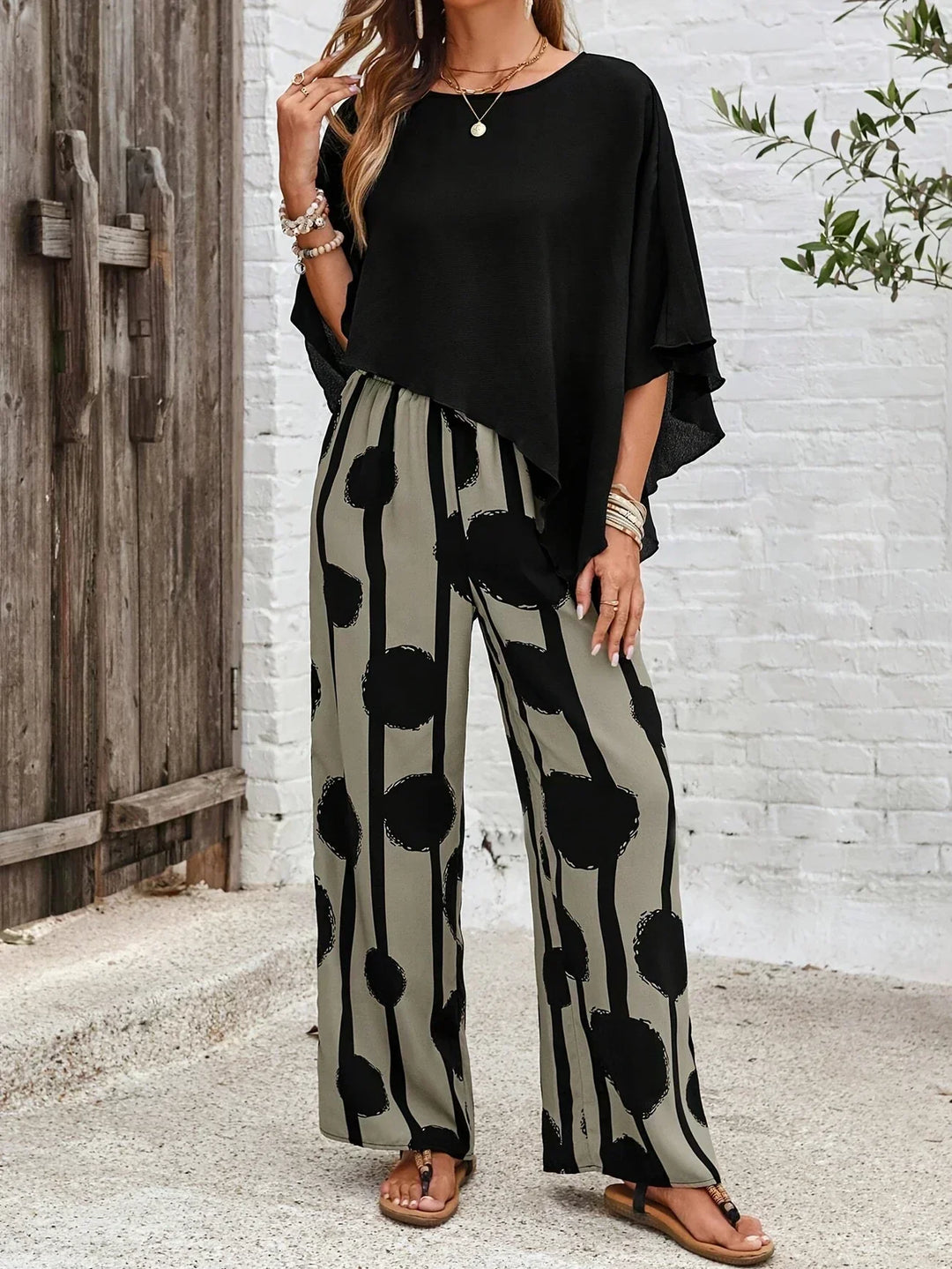 Woman wearing an asymmetrical black top with wide-leg grey pants featuring black abstract patterns, standing outdoors by a wooden gate.