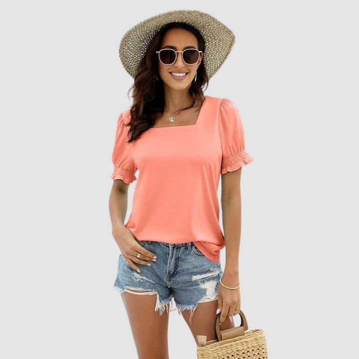 Woman styled in peach square neck top, distressed denim shorts, straw hat, carrying woven handbag in a studio shot.