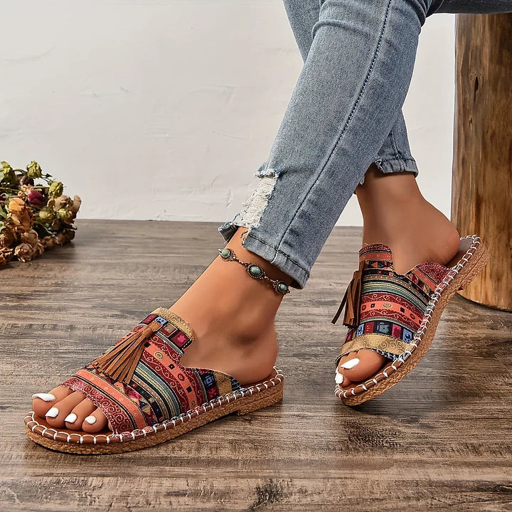 Person wearing colorful orthopedic sandals with tassels, sitting indoors on wooden floor.