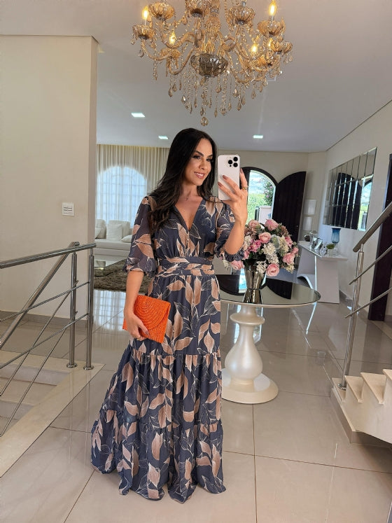 Model wearing a blue floral maxi dress with beige accents, holding a textured clutch in a chic living room setting.