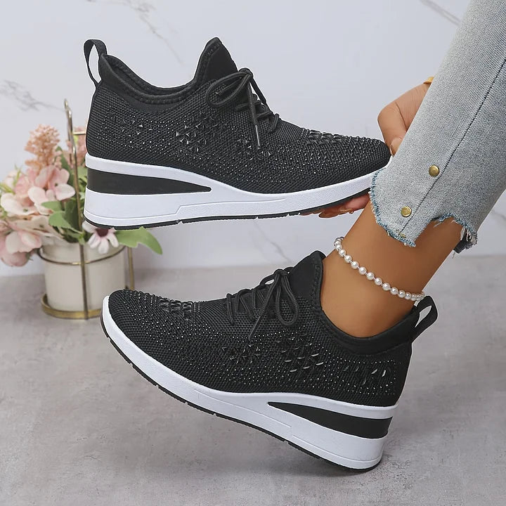 Black orthopedic sneakers with subtle shimmer worn with jeans, in a light indoor space with decorative flowers.