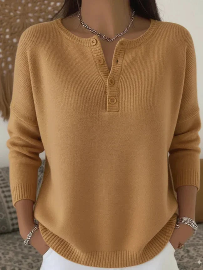 Model in tan sweater with buttoned neckline, white jeans, bracelets, indoors with a cozy background.