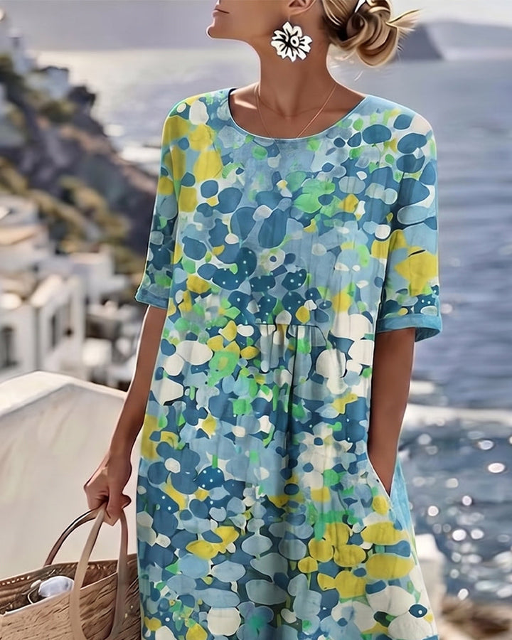 Model in blue floral midi dress on terrace, holding bag with ocean view in background.