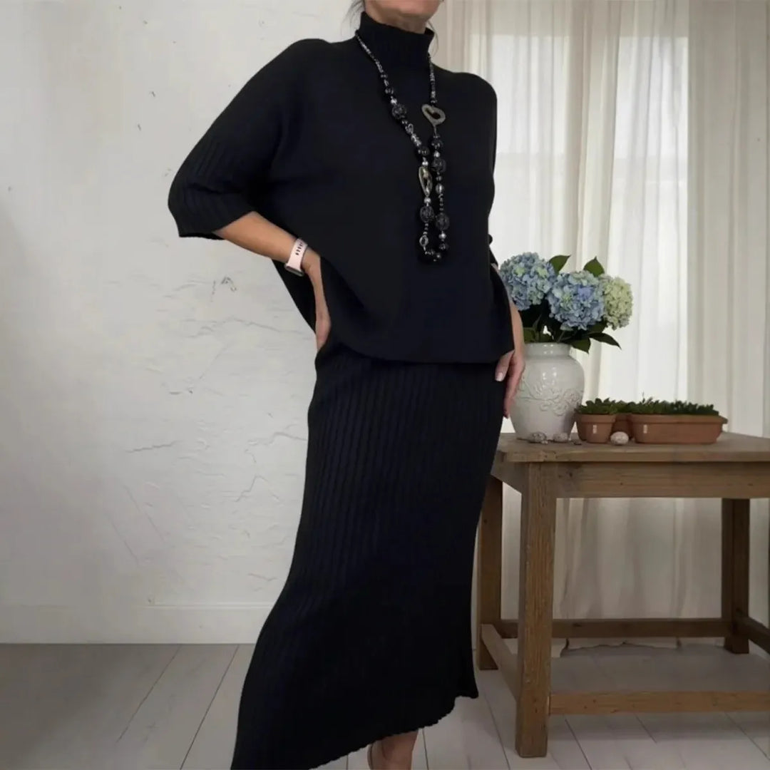 Black 2-piece set with ribbed skirt and loose top, accessorized with necklace in cozy room setting.