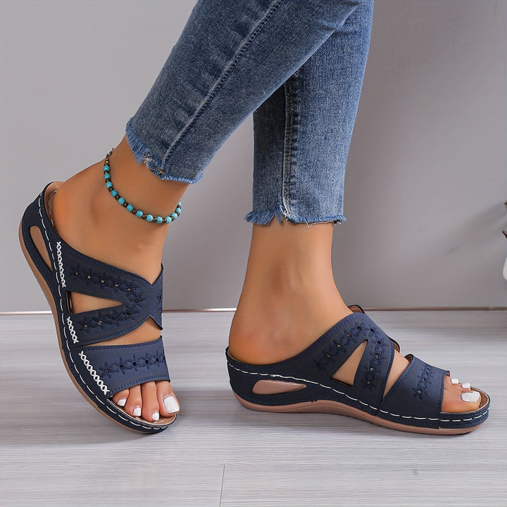 Navy orthopedic sandals with cutout design and white stitching, worn by a person in frayed jeans, on light wood floor.
