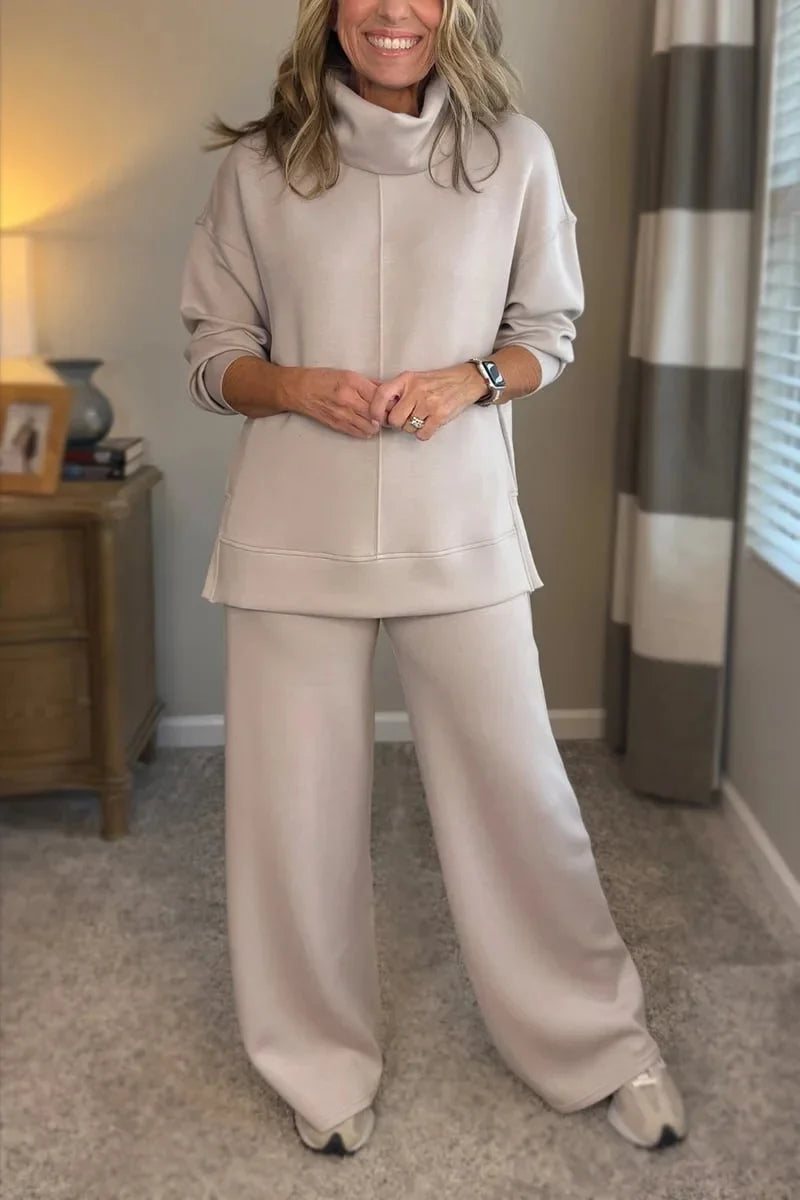 Woman in a beige comfy set with long sleeves and wide-leg pants, standing in a carpeted room with a lamp and books in the background.