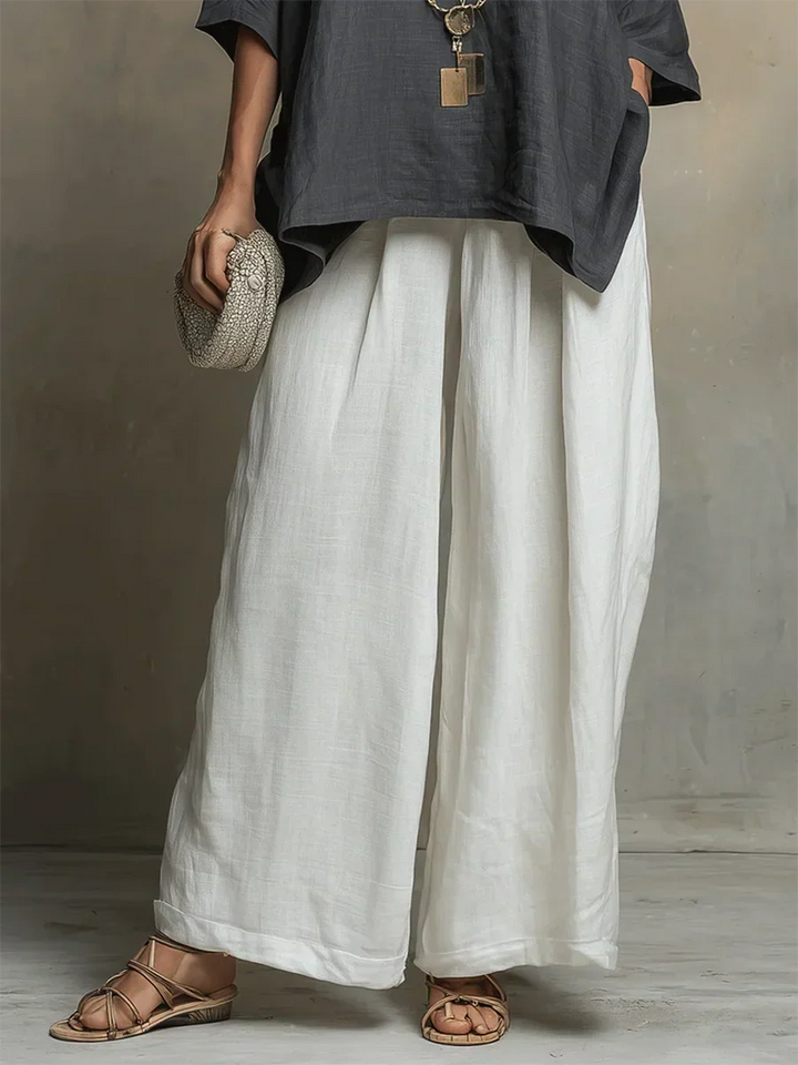Close-up of white wide-leg pants and dark top, holding textured clutch, studio backdrop.