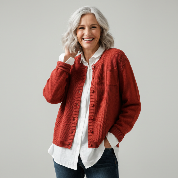 Woman in studio wearing red knit cardigan over white shirt, hand near face, smiling.