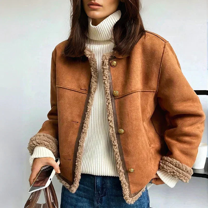 Woman wearing a cozy brown jacket with plush trim, white turtleneck, and jeans, holding a phone and bag in a bright indoor setting.