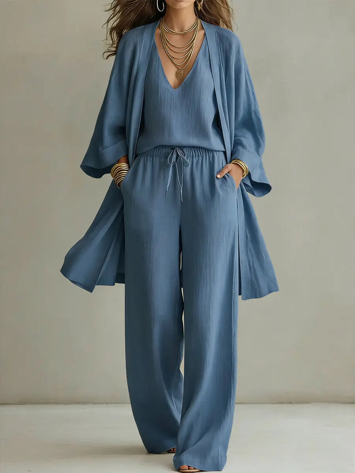 Blue three-piece relaxed homewear set with v-neck top, drawstring pants, and flowy overcoat in a studio setting.