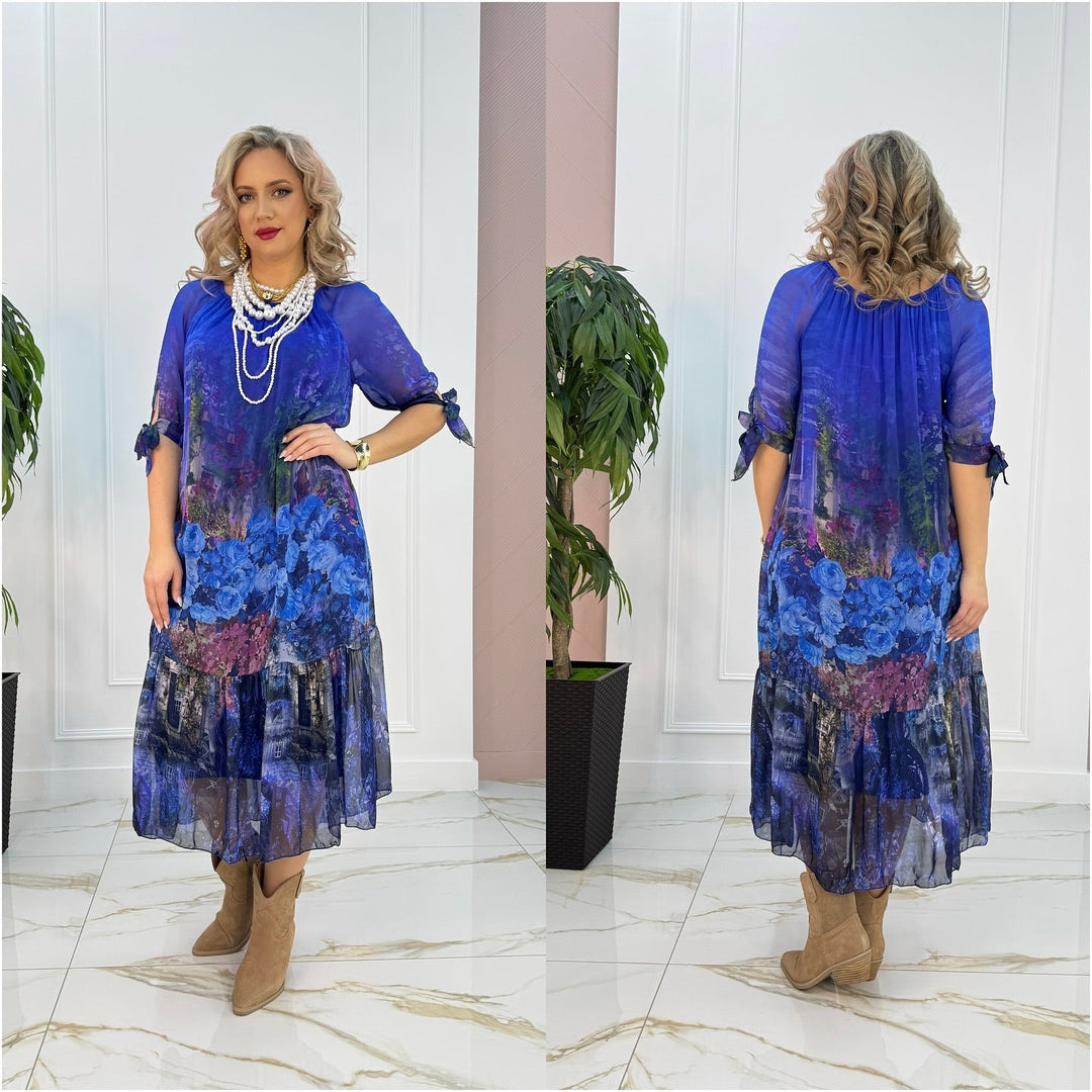 Model in blue floral maxi dress with flowing fabric, posing indoors on marble floor near plant.