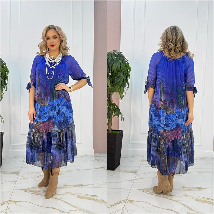 Model in blue floral maxi dress with flowing fabric, posing indoors on marble floor near plant.