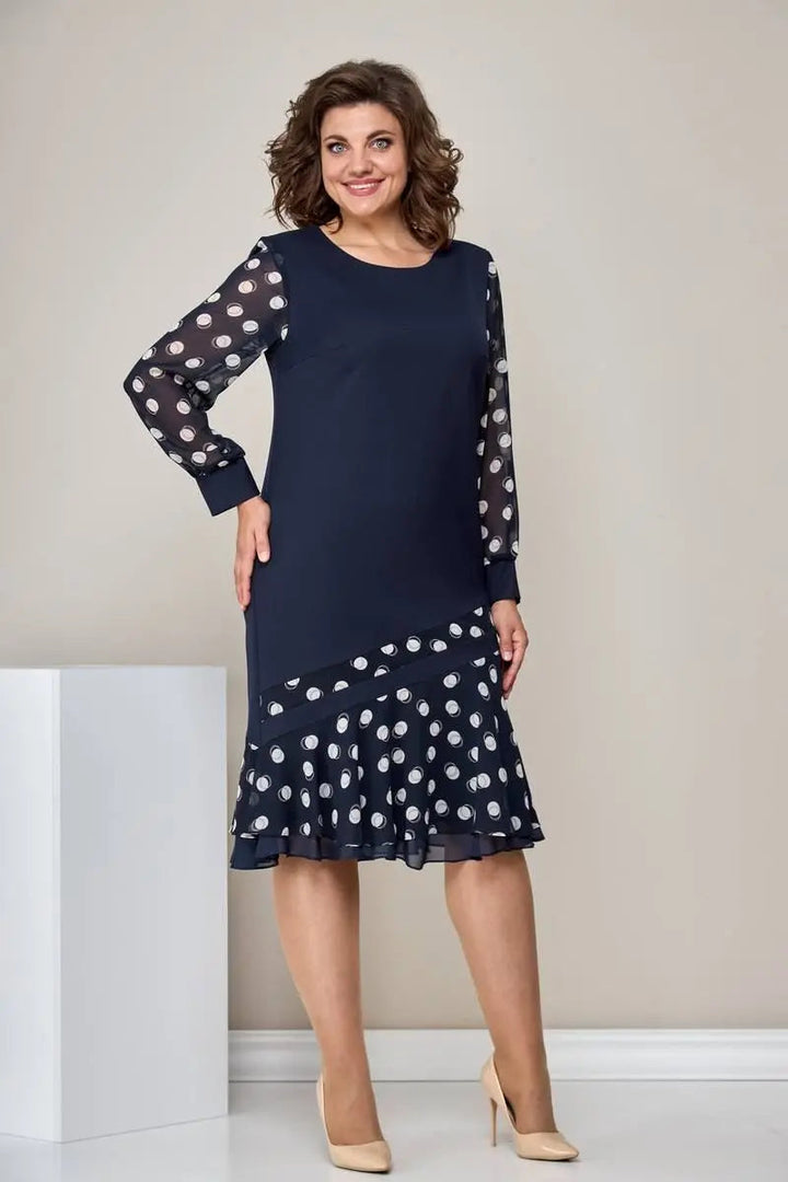Woman in navy dress with sheer polka dot sleeves and ruffled hem, beige heels, standing in a neutral room.