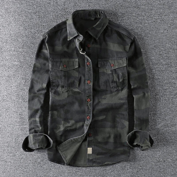 Dark urban camo shirt laid out on a gray surface, showcasing front pockets and folded sleeves.