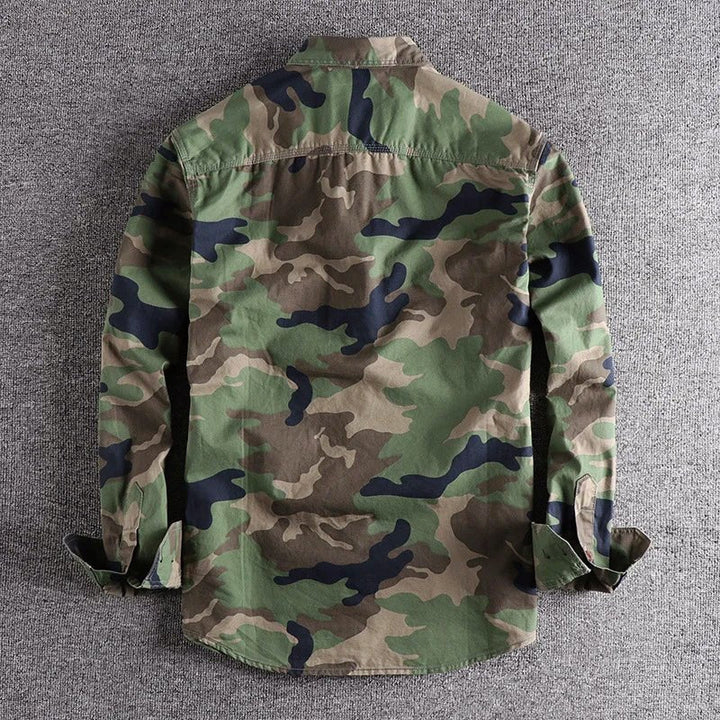 Back view of green urban camo shirt lying on a gray surface, with sleeves softly folded.