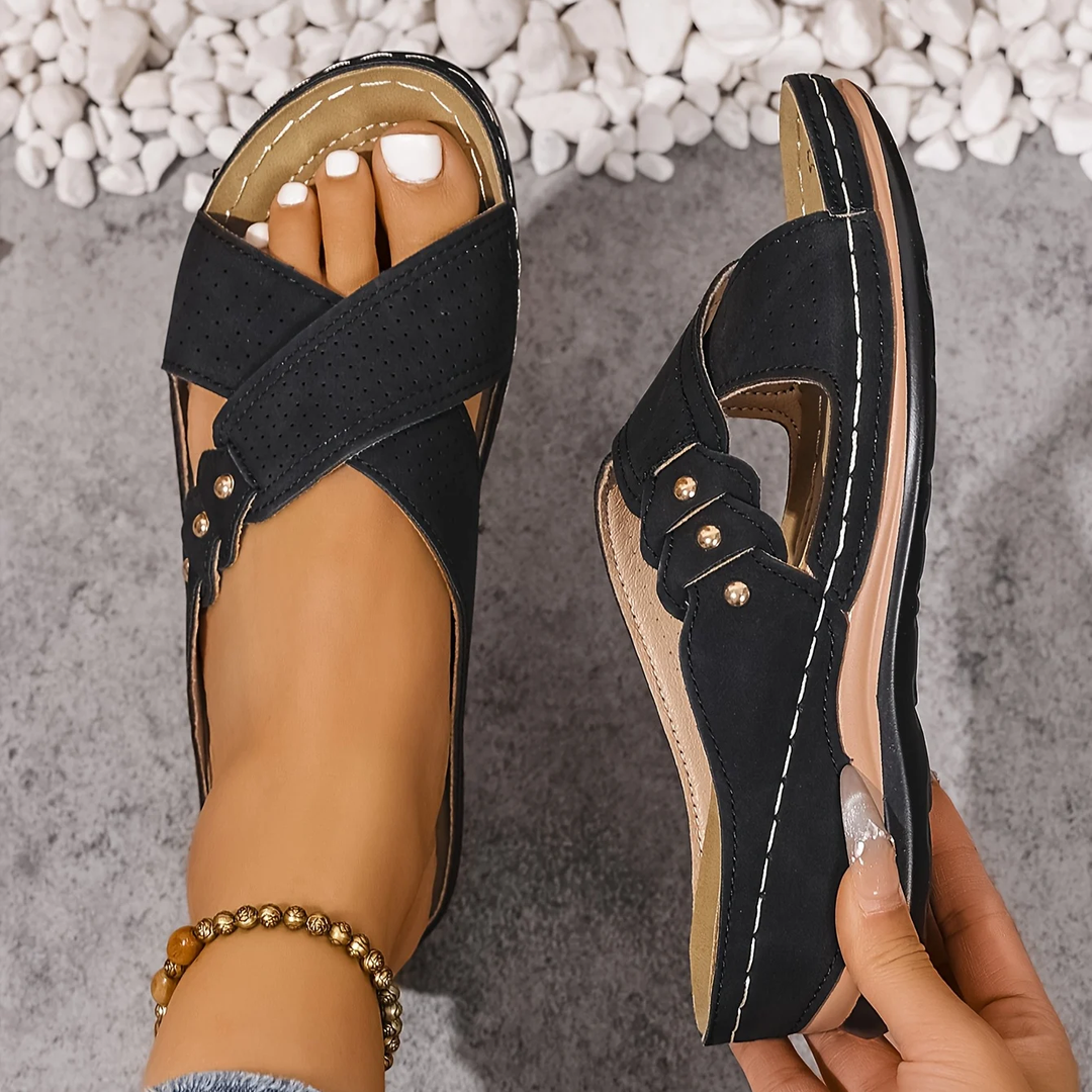 Black orthopedic wedge sandals held in hand, shown on a textured surface with white stones.
